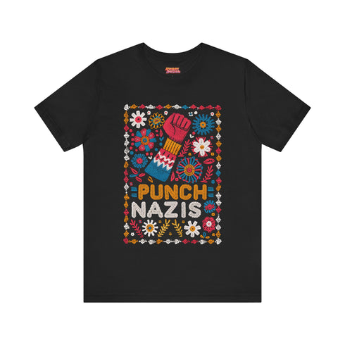 Black t-shirt featuring an embroidered-style design with the phrase "PUNCH NAZIS" in bold, colorful lettering. Above the text, a raised red fist is illustrated, symbolizing resistance, and is surrounded by a vibrant folk-art-inspired floral border with bright flowers, leaves, and geometric patterns in red, yellow, blue, and white.